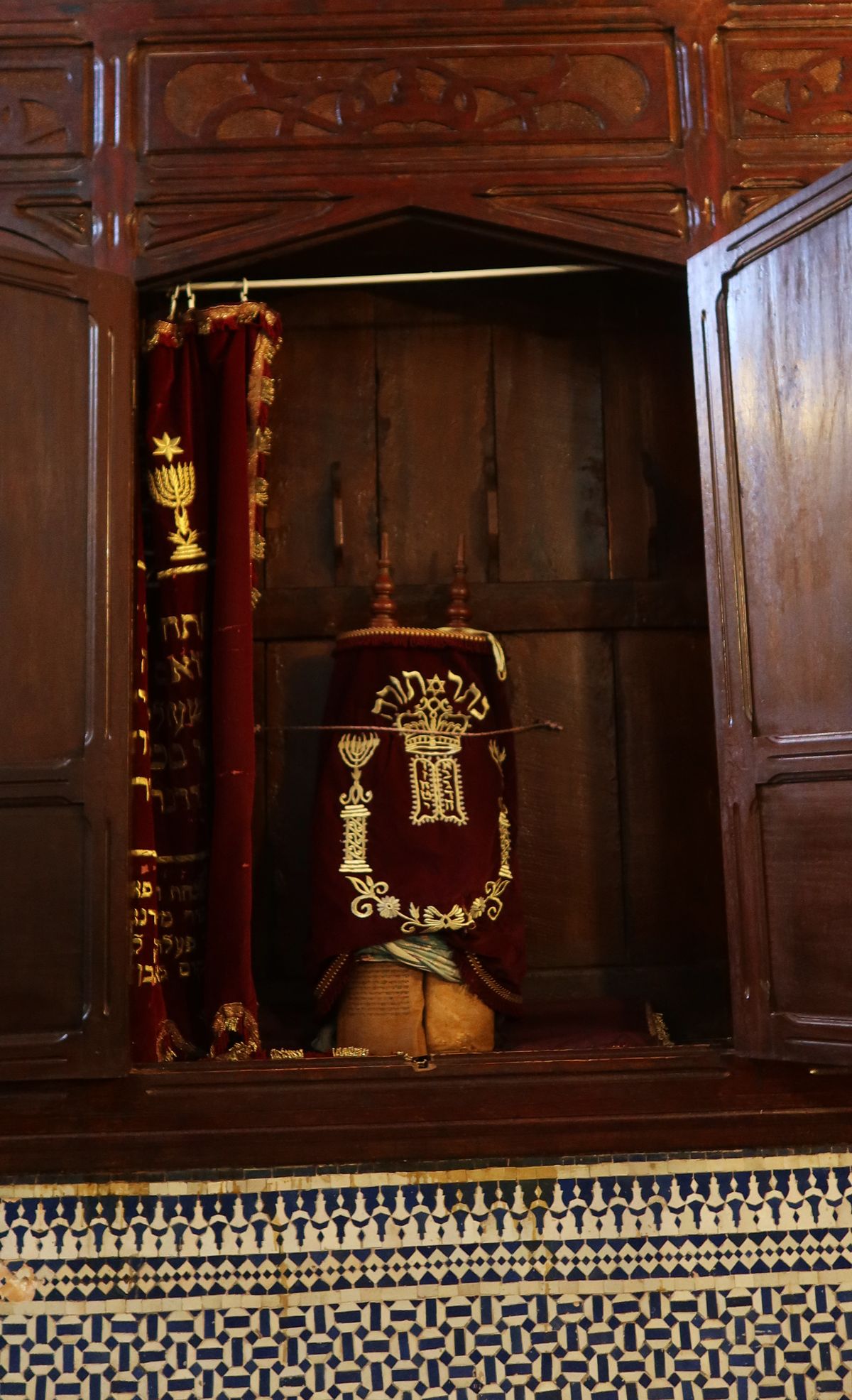 A Torah scroll in its velvet mantle and silver crown in the ark of a Moroccan synagogue — the zellige tilework below the wooden ark is characteristic of the Andalusian-Moroccan craft tradition applied to Jewish religious spaces